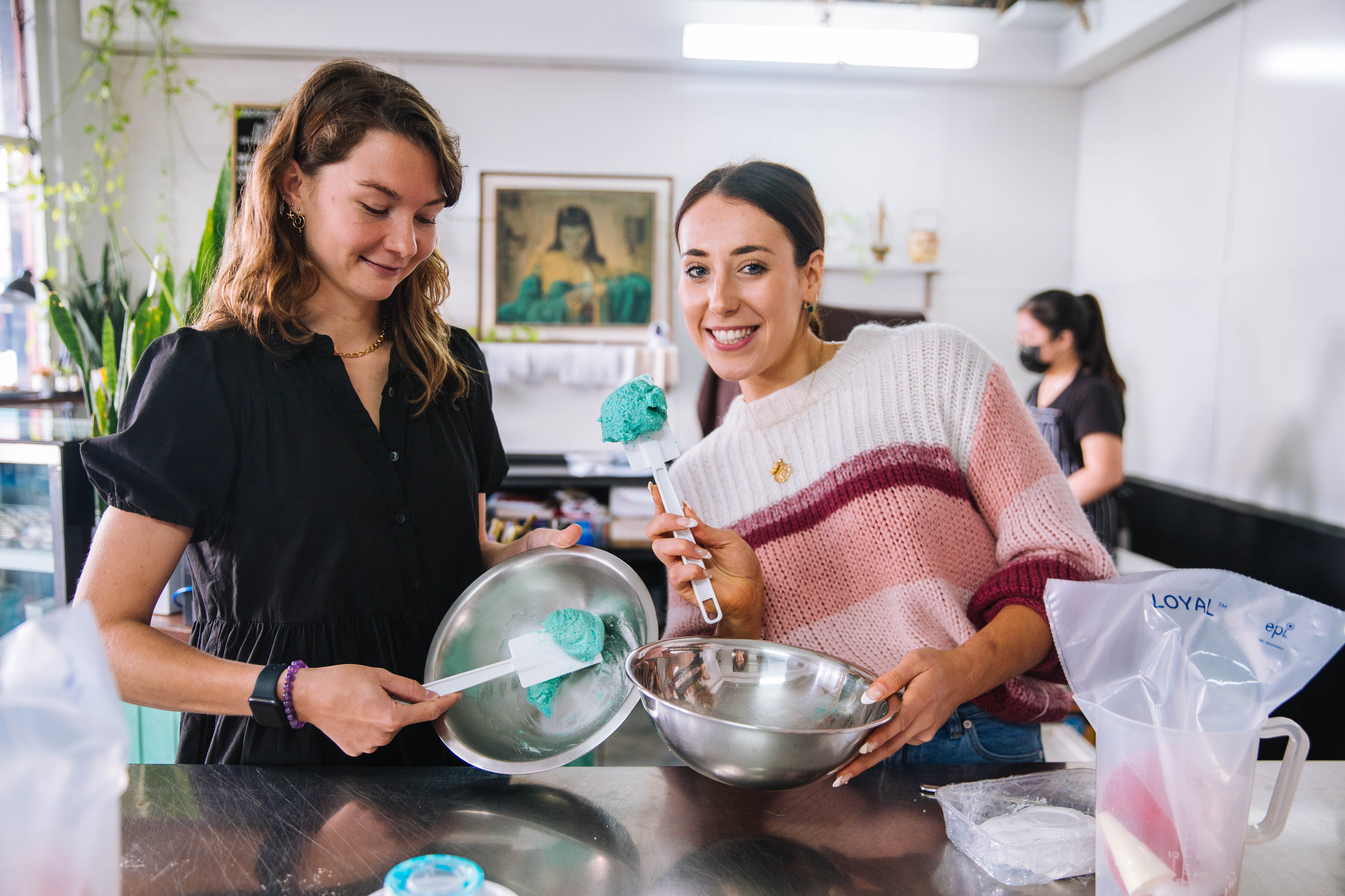 Two smiling women holding silver mixing bowls containing blue batter
